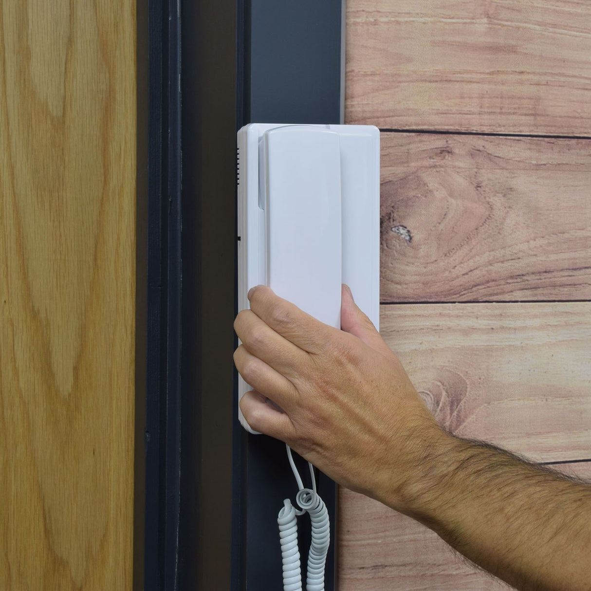 Hand lifting the white Mercury 2-Way Intercom handset from its wall-mounted base beside a wood-panelled door, coiled cable visible below.