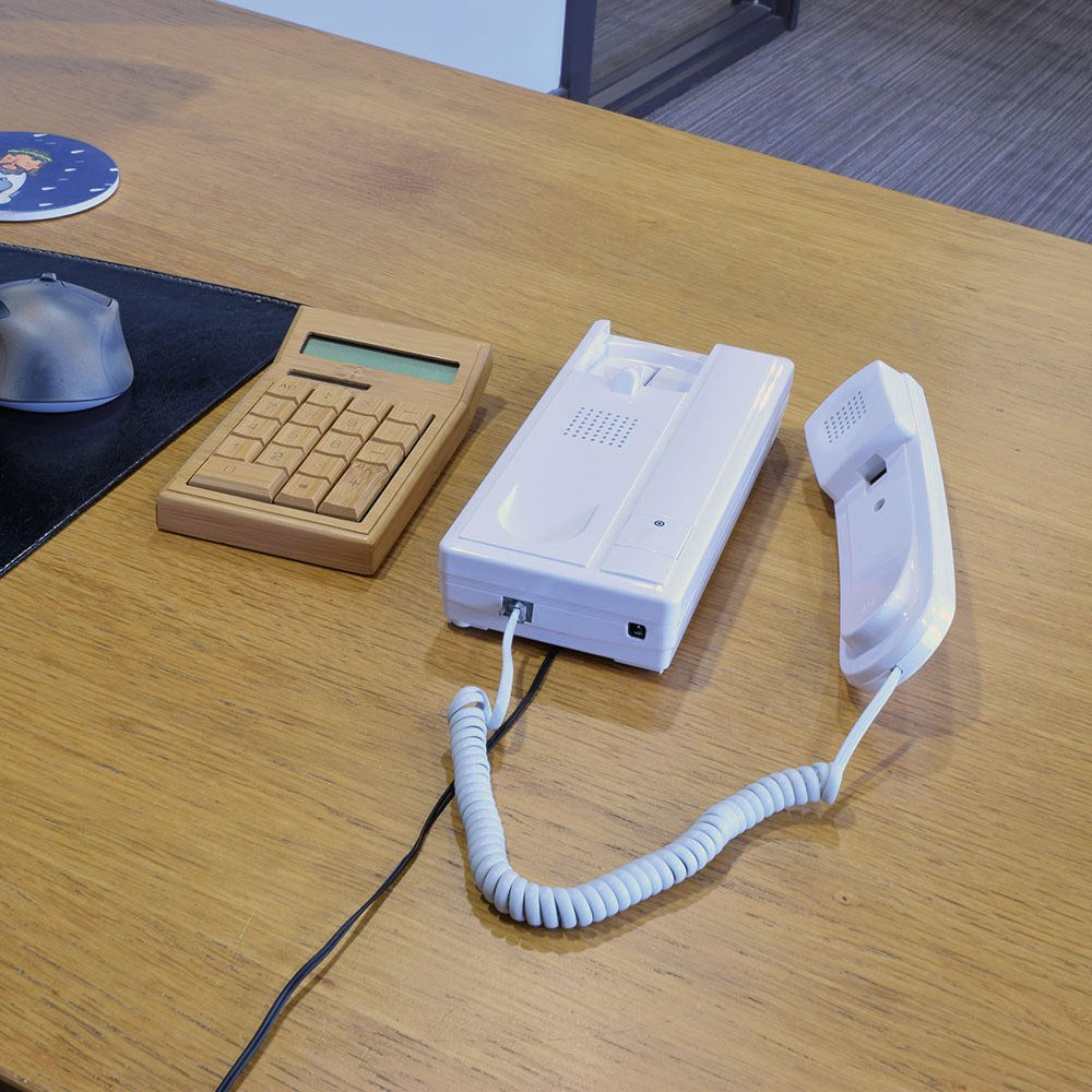 AVSL Mercury 2-Way Intercom wired audio unit in white plastic with lifted handset and coiled cord, shown on a wooden desk beside a calculator and mouse.