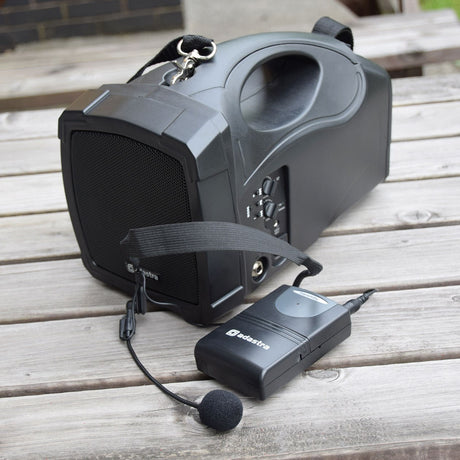 Adastra VH174.8 VHF portable PA speaker with carry strap alongside a black bodypack transmitter and headset microphone, shown outdoors on a wooden table.