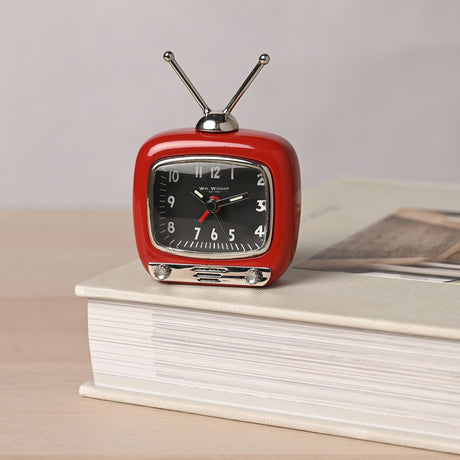 Miniature television-shaped alarm clock in glossy red with twin silver aerials, chrome bezel, black dial with silver numerals, and a base panel with knobs, shown resting on a book.