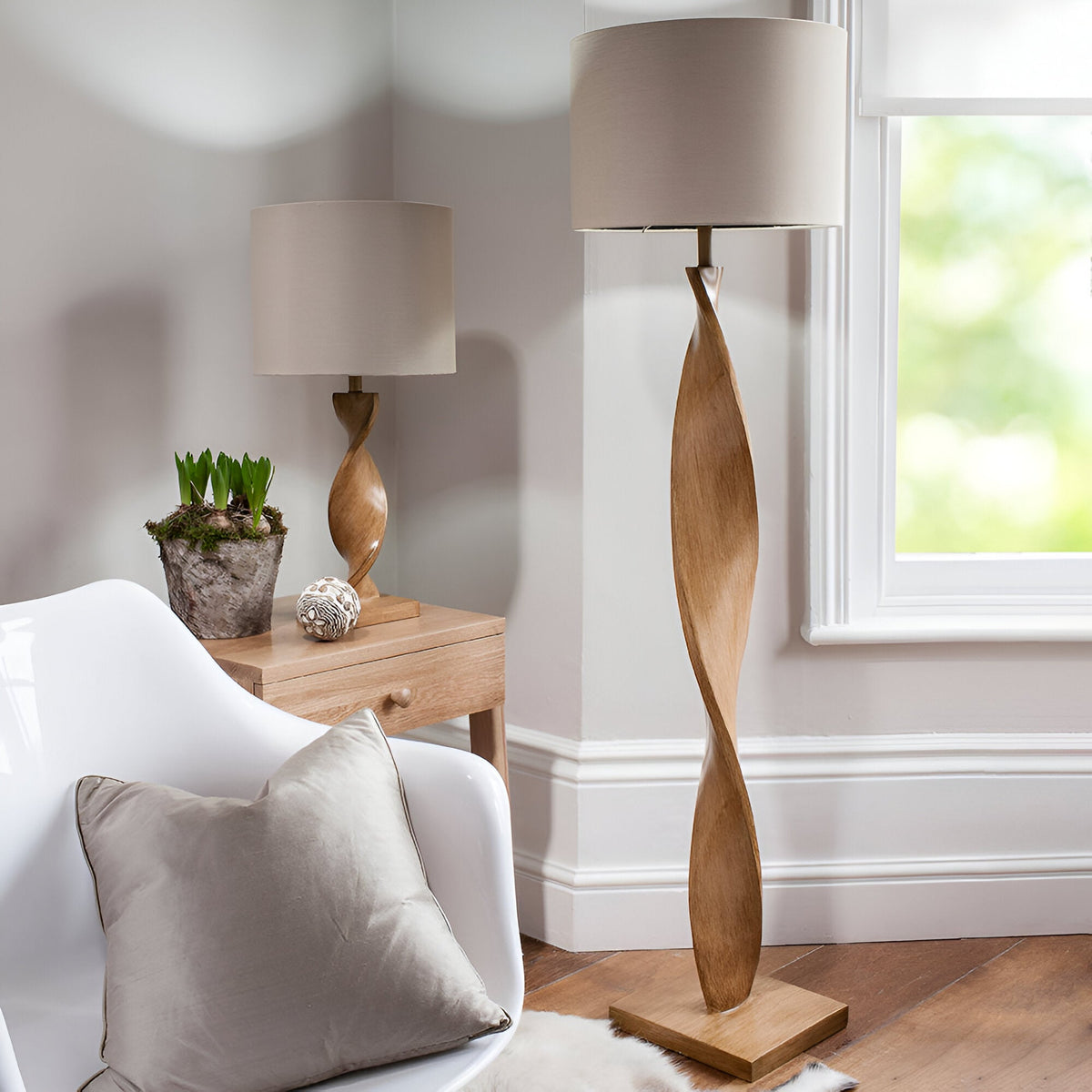 Alcott Table Lamp in Natural on a wood side table, showing its twisted oak-effect spiral base and cylindrical linen shade in a living room setting.