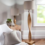 Alcott Table Lamp in Natural on a wood side table, showing its twisted oak-effect spiral base and cylindrical linen shade in a living room setting.