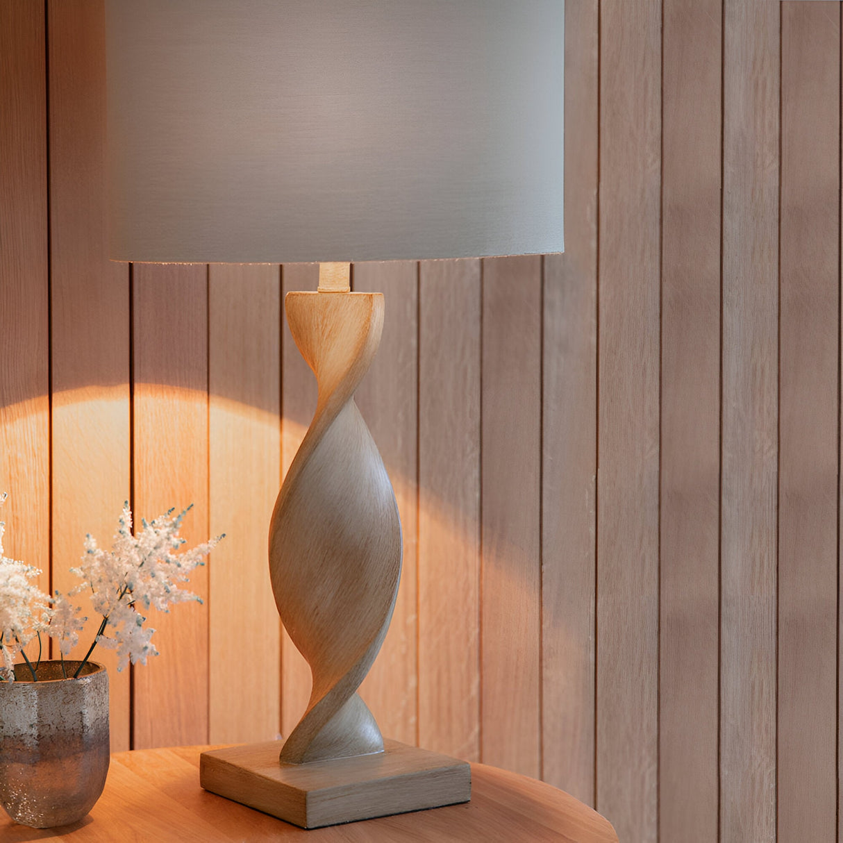 Alcott Table Lamp in Natural with twisted light oak-effect wood base and grey linen cylinder shade, lit on a wooden table against wood-panelled walls.
