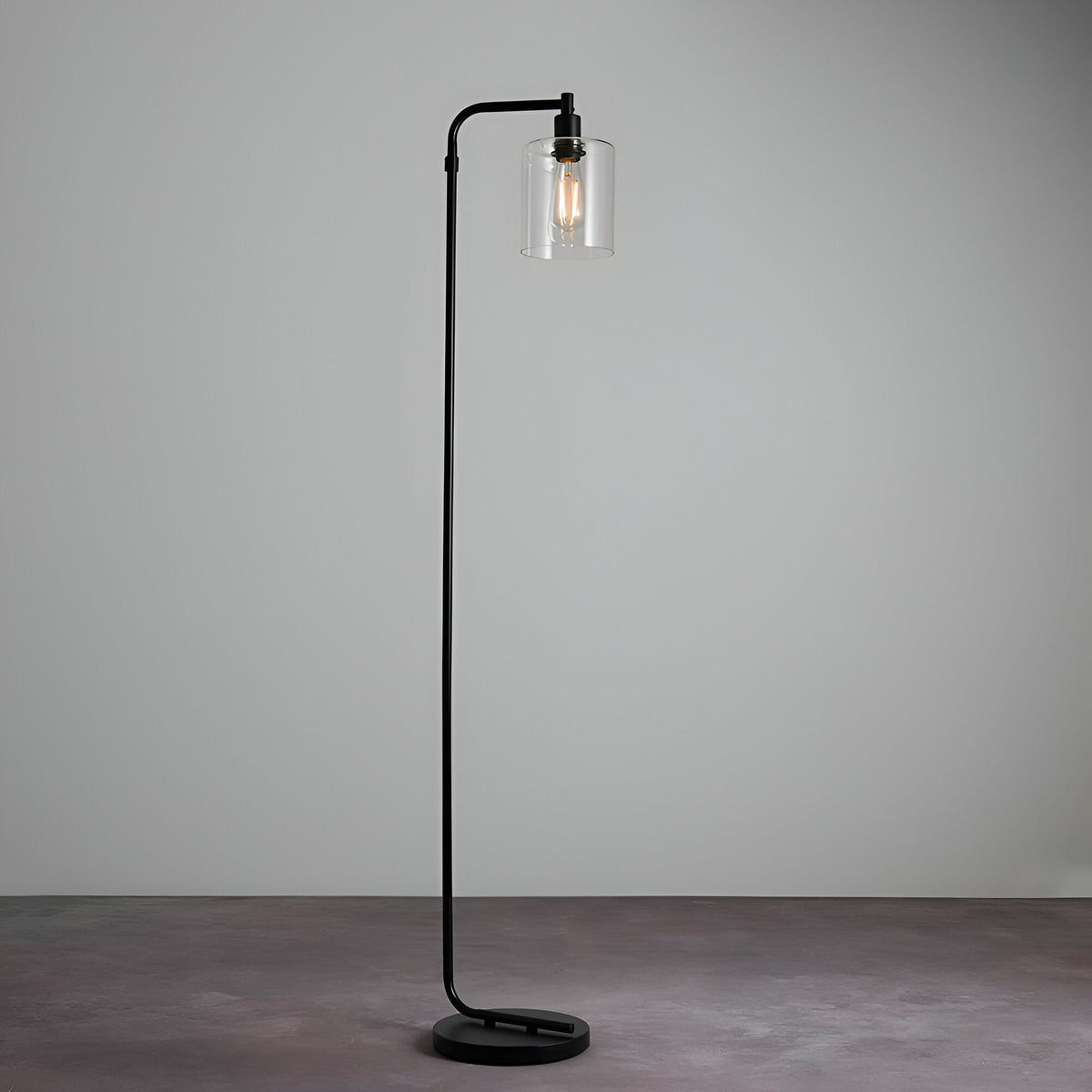 Cranmere Floor Lamp in matt black metal with a curved arm and clear glass shade over an exposed bulb, on a round base, set against a plain grey background.
