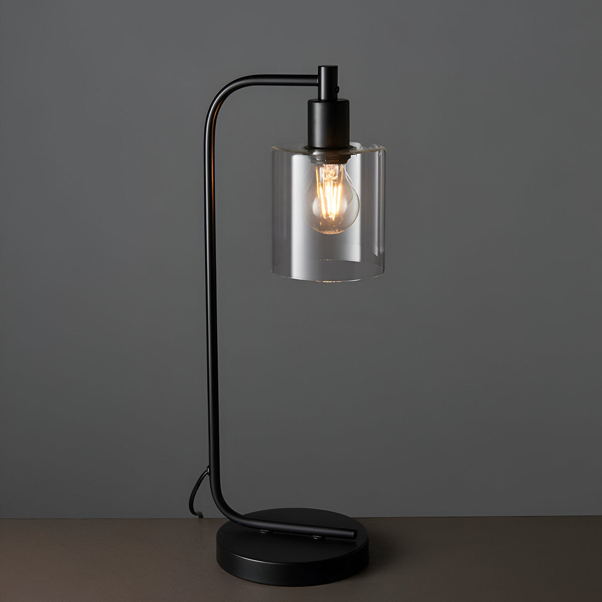 Endon Cranmere table lamp in matt black metal with a clear glass cylinder shade and exposed filament bulb, shown lit against a grey background.