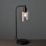 Endon Cranmere table lamp in matt black metal with a clear glass cylinder shade and exposed filament bulb, shown lit against a grey background.