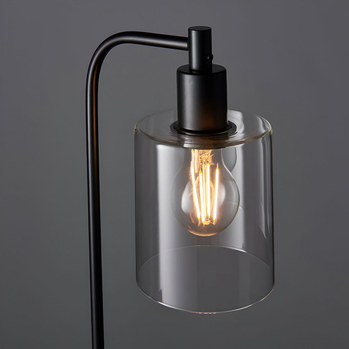 Endon Cranmere table lamp in matt black with arched metal arm and clear glass cylinder shade housing a lit E27 filament bulb, on a grey background.