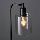 Endon Cranmere table lamp in matt black with arched metal arm and clear glass cylinder shade housing a lit E27 filament bulb, on a grey background.