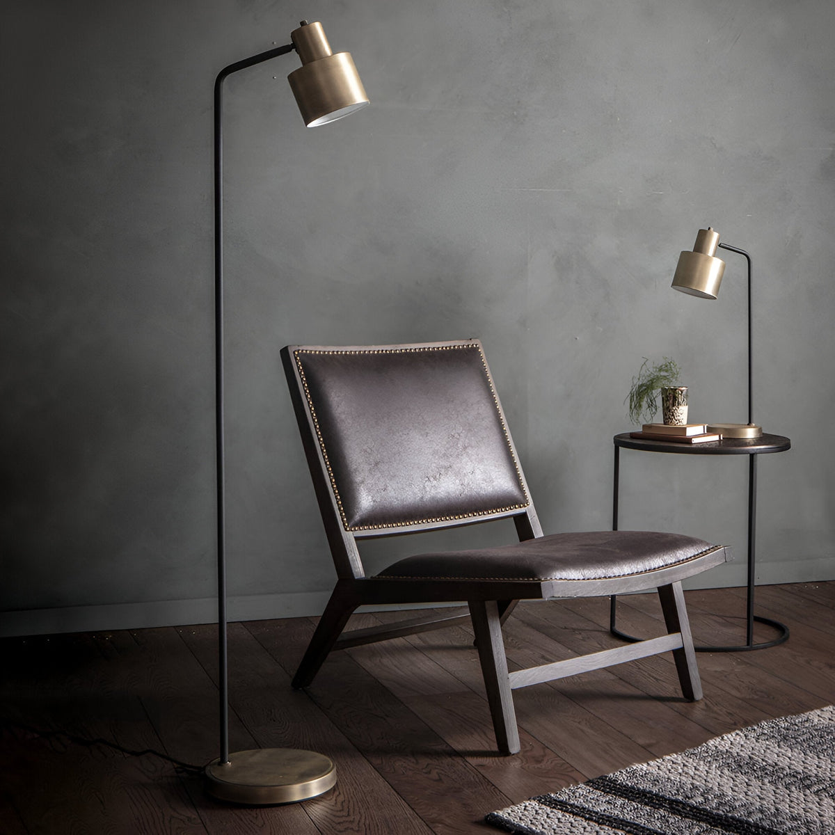 Carlbourne Table Lamp in matt black and antique brass, industrial-style with a curved arm and cylindrical shade, shown on a side table beside an armchair.