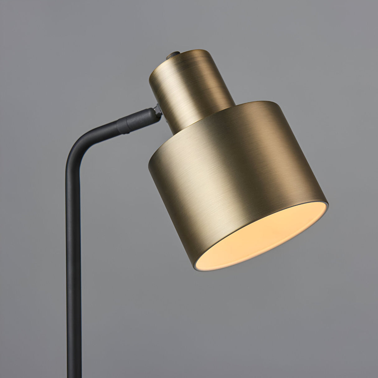 Close-up of the Carlbourne Table Lamp's cylindrical shade in matt antique brass, angled on a black rod and joint, against a plain grey background.