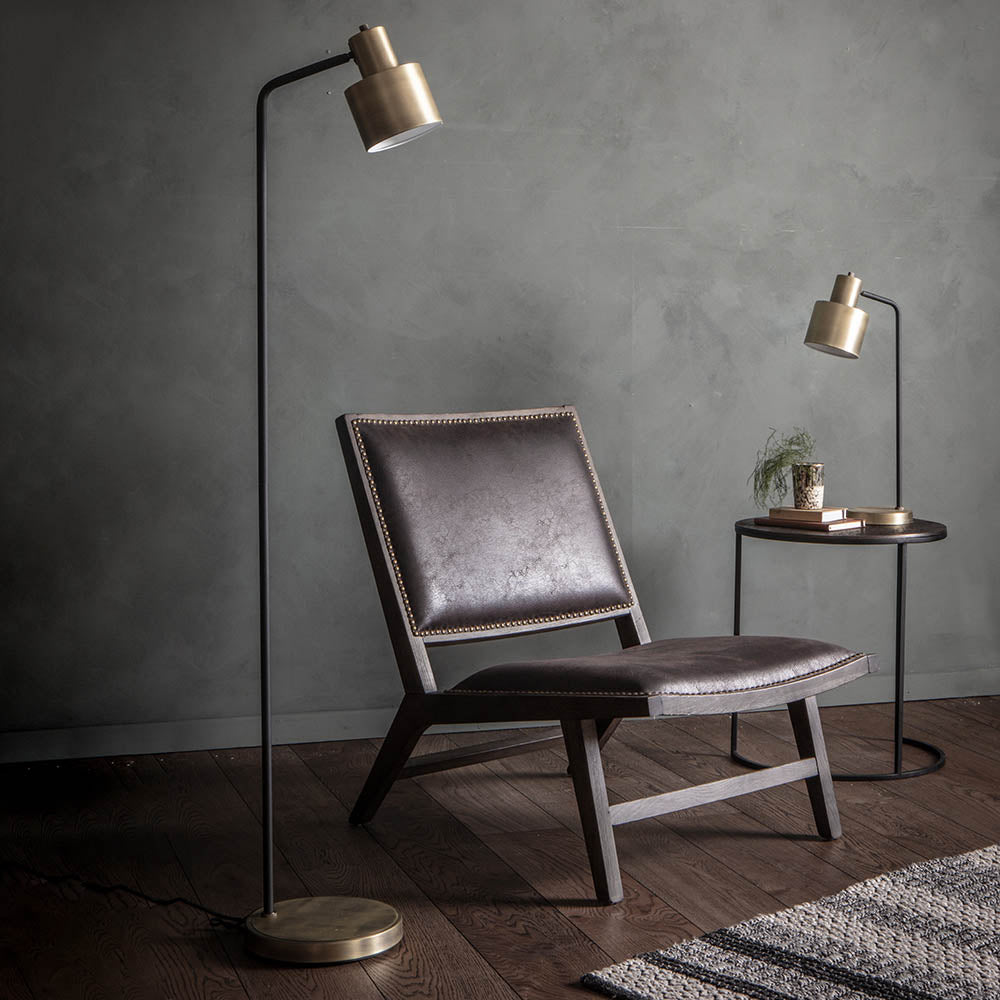Carlbourne Floor Lamp in brass and black with an angled arm and cylindrical shade, shown beside a leather chair and side table.