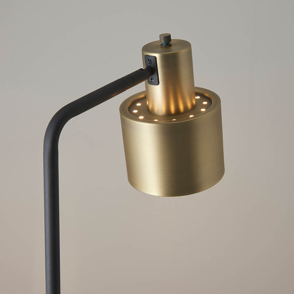 Close-up of the Carlbourne Floor Lamp's brass cylindrical head with perforated top and knurled dial, on a black bent arm, against a plain grey background.
