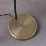 Close-up of the Carlbourne Floor Lamp's round brass base with black metal pole and cable, shown on a grey surface.