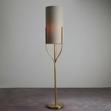 Endon Bramdon floor lamp in satin brass with a natural linen cylinder shade on a slender stem with curved supports, shown on a neutral background.