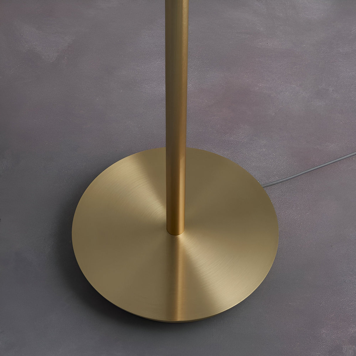 Close-up of the Endon Bramdon floor lamp base, showing the round satin brass plate and cylindrical stem on a grey floor with grey fabric cable.