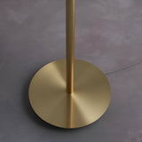 Close-up of the Endon Bramdon floor lamp base, showing the round satin brass plate and cylindrical stem on a grey floor with grey fabric cable.