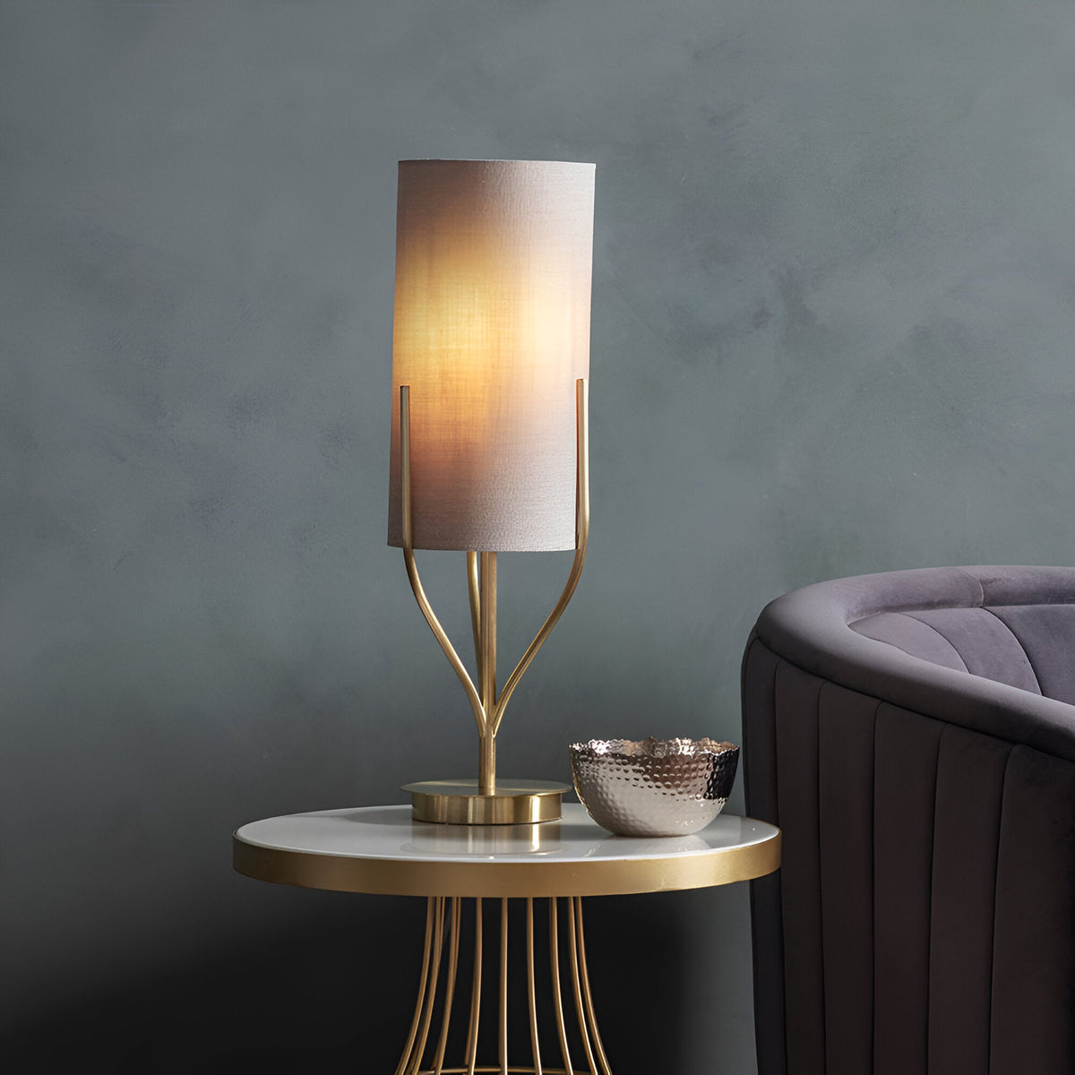 Endon Bramdon table lamp in satin brass with a natural linen cylinder shade, lit on a round gold side table beside a grey velvet armchair.