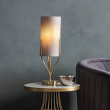 Endon Bramdon table lamp in satin brass with a natural linen cylinder shade, lit on a round gold side table beside a grey velvet armchair.