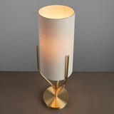 Endon Bramdon table lamp with satin brass base, upright prongs, and tall natural linen cylinder shade, illuminated against a light grey backdrop.