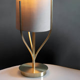 Endon Bramdon table lamp close-up showing satin brass branching stem, round base and natural linen cylinder shade on a pale surface against grey.