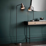 Endon Carlbourne floor lamp in brushed silver and matt black, lit beside a wooden console with matching table lamp against a dark teal wall.