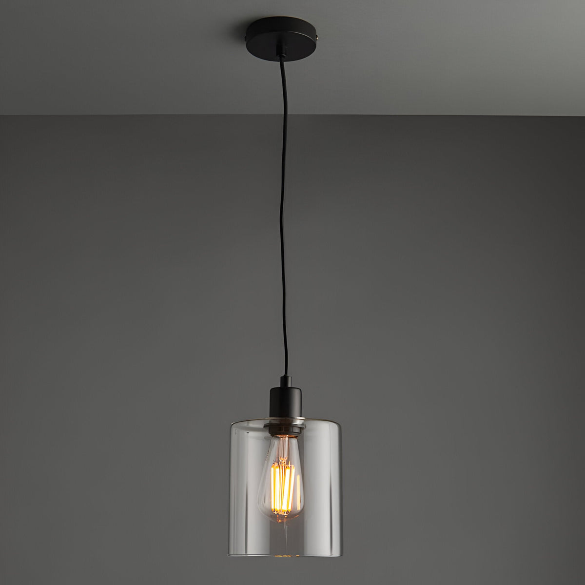 Endon Cranmere pendant light in matt black with a compact clear glass cylinder shade and exposed filament bulb on a black flex against a grey ceiling.