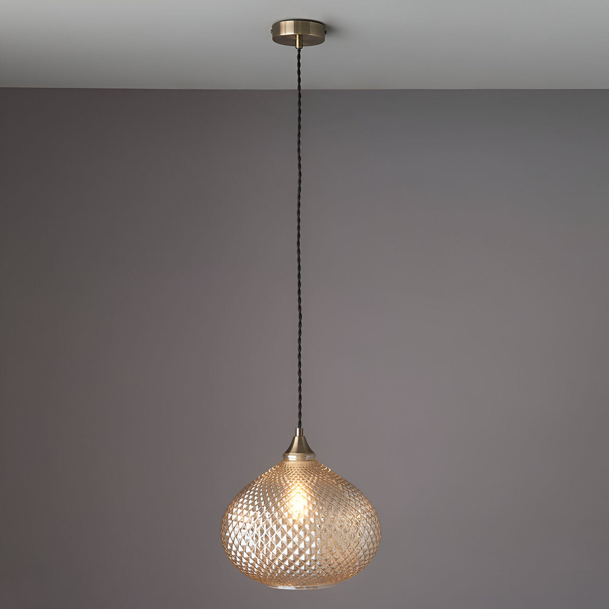 Endon Caldstead pendant light with champagne glass teardrop shade and antique brass fitting on a black flex, shown lit against a grey wall.