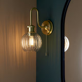 Endon Brockshaw satin brass bathroom wall light with swan-neck arm, ribbed clear glass globe shade and pull switch, mounted beside a black-framed mirror.