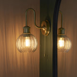Endon Brockshaw satin brass bathroom wall light with swan-neck arm and clear ribbed glass globe shade, shown illuminated beside a mirror reflection.