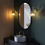 Endon Brockshaw satin brass bathroom wall lights with ribbed glass shades flanking an oval black-framed mirror above a vanity sink in a dark green bathroom.