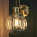 Endon Brockshaw bathroom wall light in satin brass with swan-neck arm, knurled detail and clear ribbed glass globe shade around a lit E27 bulb.