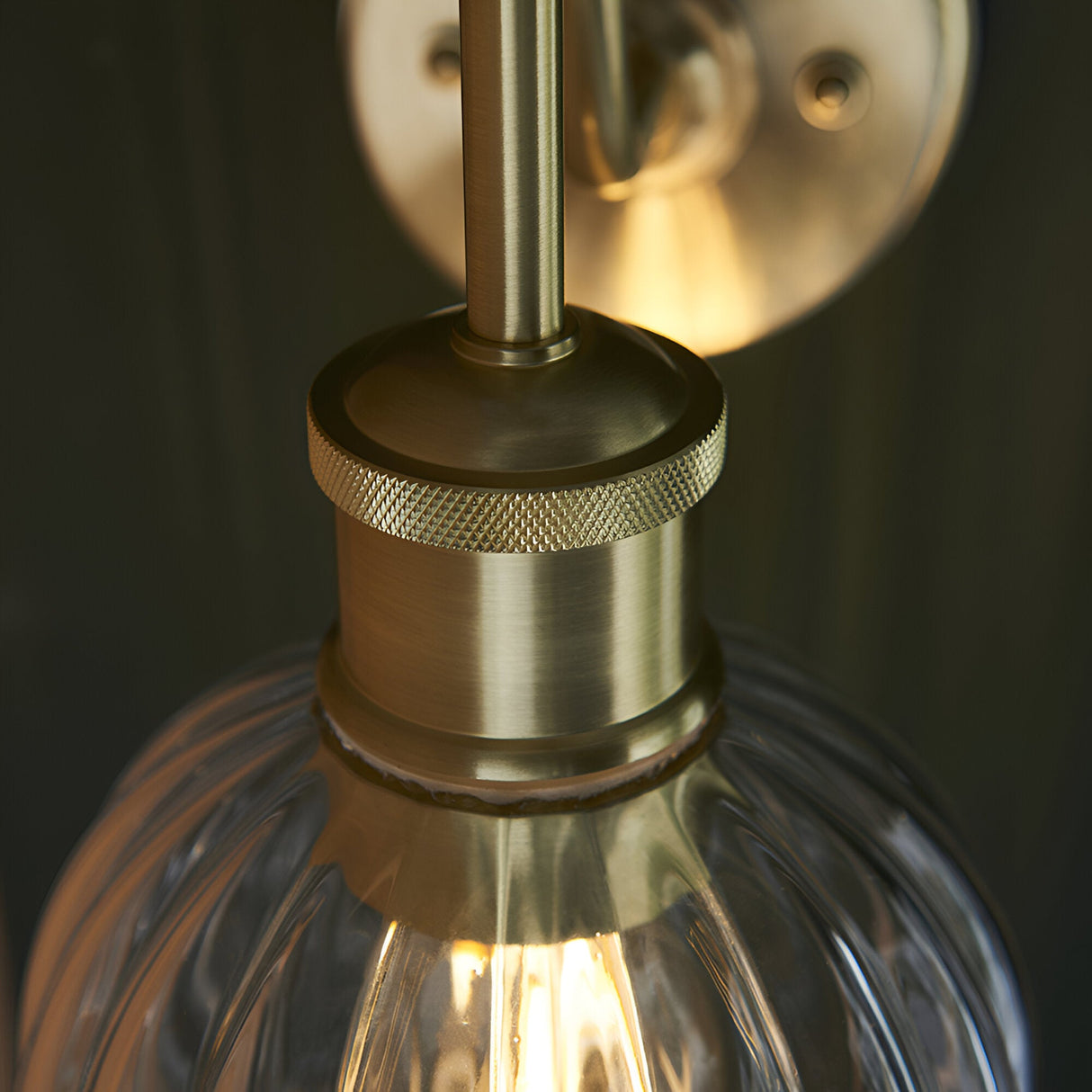 Close-up of Endon Brockshaw bathroom wall light in satin brass, showing knurled lamp holder detail and clear ribbed glass shade with lit E27 bulb.