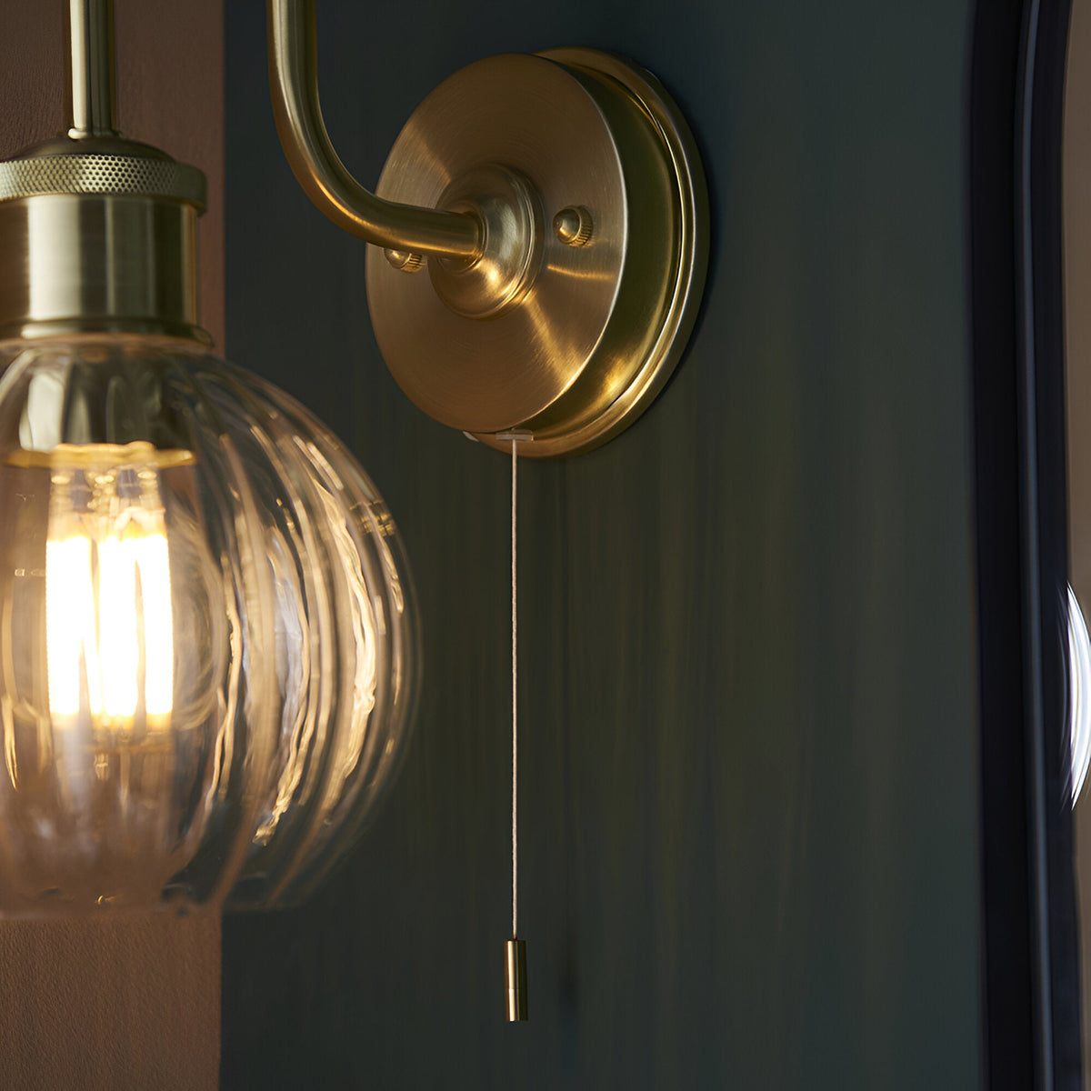 Endon Brockshaw bathroom wall light in satin brass with swan-neck arm, knurled detail, ribbed glass shade, pull switch and lit E27 bulb.