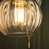 Close-up of Endon Brockshaw bathroom wall light showing clear ribbed glass globe shade illuminated by an E27 filament bulb, with satin brass pull switch below.