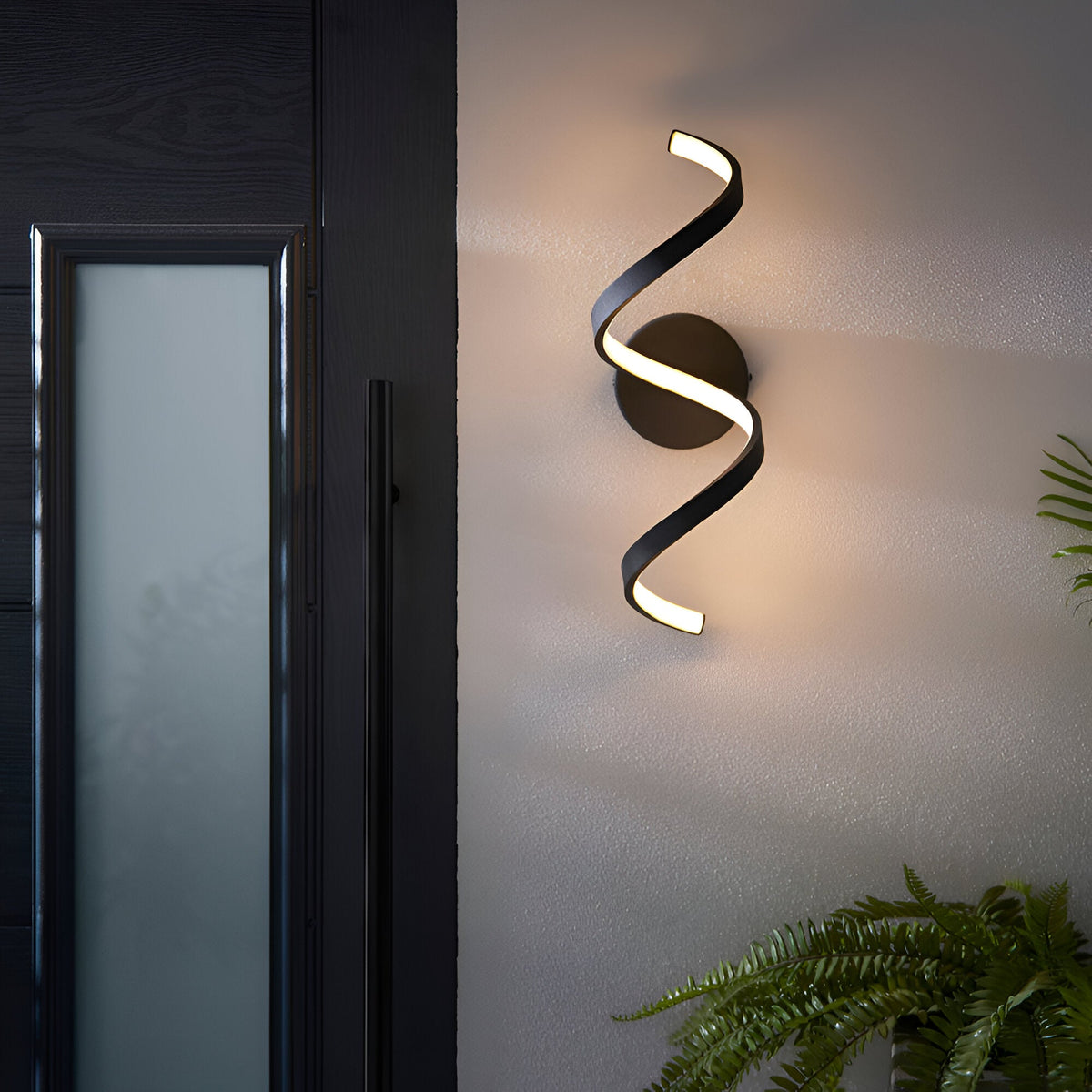 Endon Aldleigh LED wall light in black with a tall twisting spiral form, illuminated warm white beside a dark glazed entrance door.