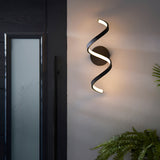 Endon Aldleigh LED wall light in black with a tall twisting spiral form, illuminated warm white beside a dark glazed entrance door.