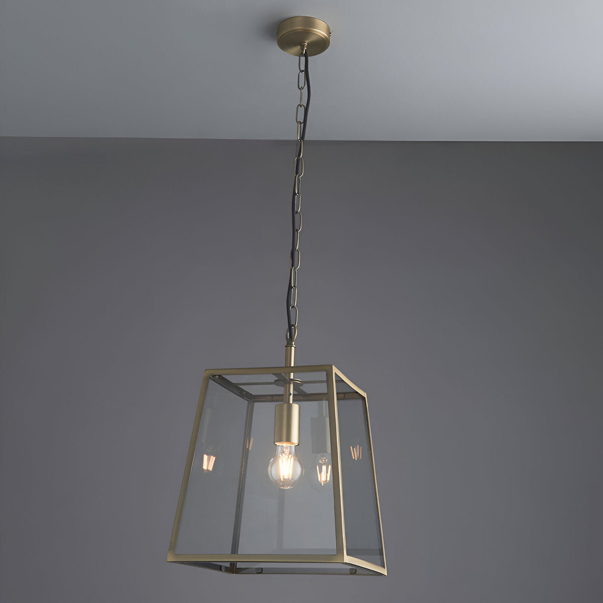 Endon Brenton lantern pendant in brass with tapered metal frame, clear glass panels and filament bulb, suspended from a chain against a grey ceiling.