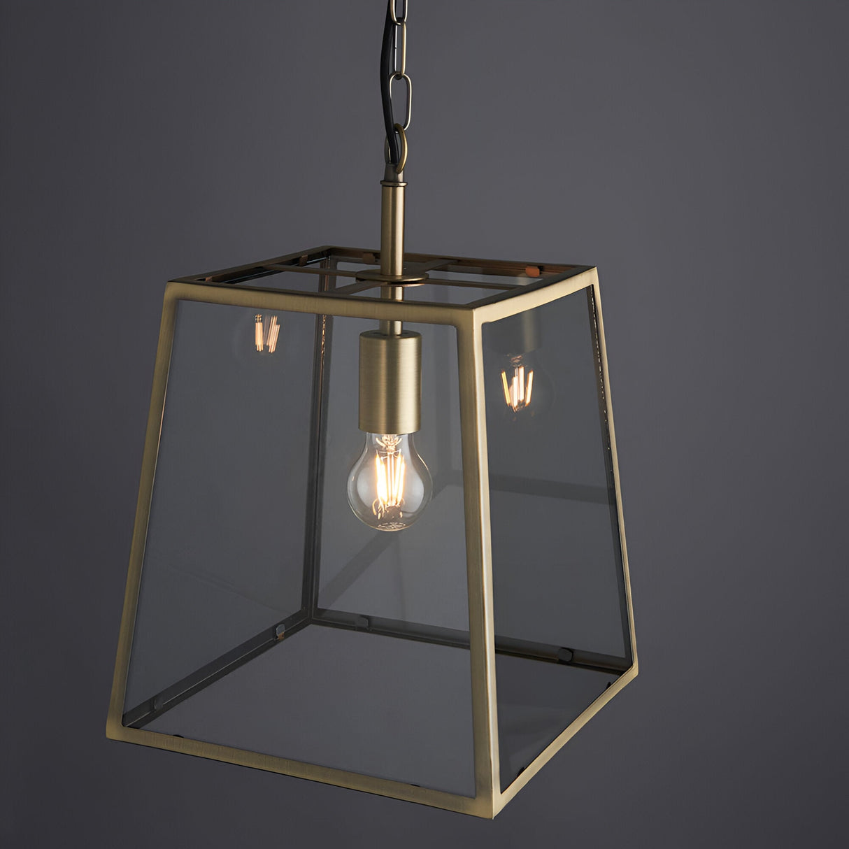 Endon Brenton tapered box lantern pendant in brass with clear glass panels, single E27 filament bulb, and black chain suspension on a grey background.