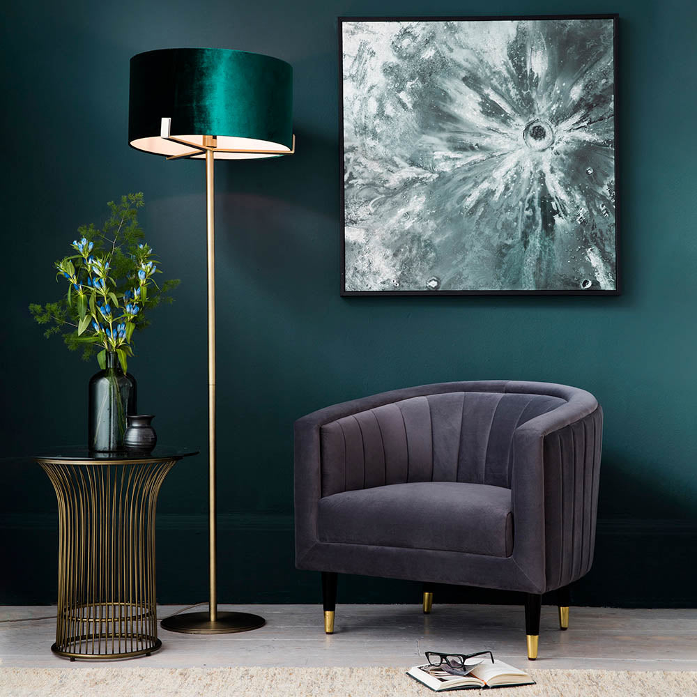 Endon Brendon floor lamp with slim antique brass stem and green velvet cylinder shade, beside a grey velvet armchair and abstract artwork.