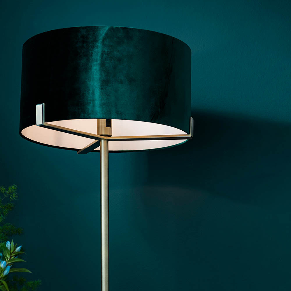 Endon Brendon floor lamp with antique brass metalwork and a green velvet cylinder shade seated on the frame, shown against a deep teal wall.