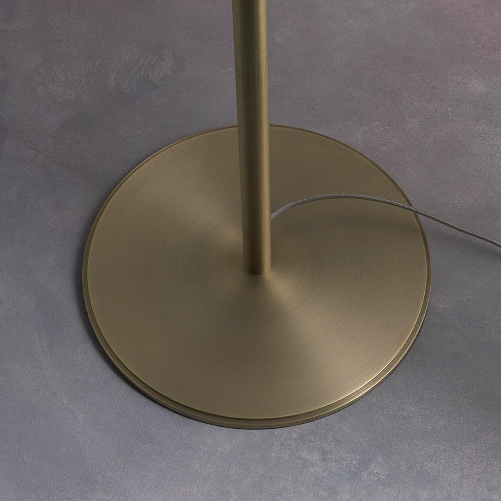 Close-up of the Endon Brendon floor lamp's circular antique brass base and slim stem, with grey fabric cable trailing across a light grey floor.