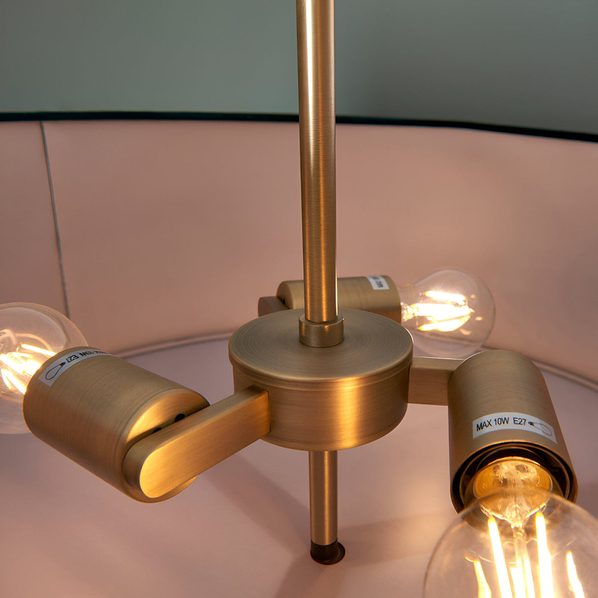 Close-up of the Brendon 3 Light Pendant's brass fitting with three angled arms holding exposed E27 bulbs beneath a pale pink fabric shade.