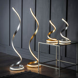 Endon Neve floor and table LED lamps with spiralling silver leaf curves and white diffusers, displayed beside a glass side table against grey panelling.