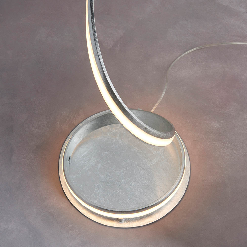 Endon Neve LED floor lamp base with silver leaf curved arm and illuminated white diffuser resting on a round disc, shown on a light grey floor.