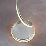 Endon Neve LED floor lamp base with silver leaf curved arm and illuminated white diffuser resting on a round disc, shown on a light grey floor.