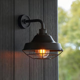 Caldton Outdoor Wall Light in matt black, industrial style with wire cage shade and lit filament bulb, mounted on a wooden panelled wall.