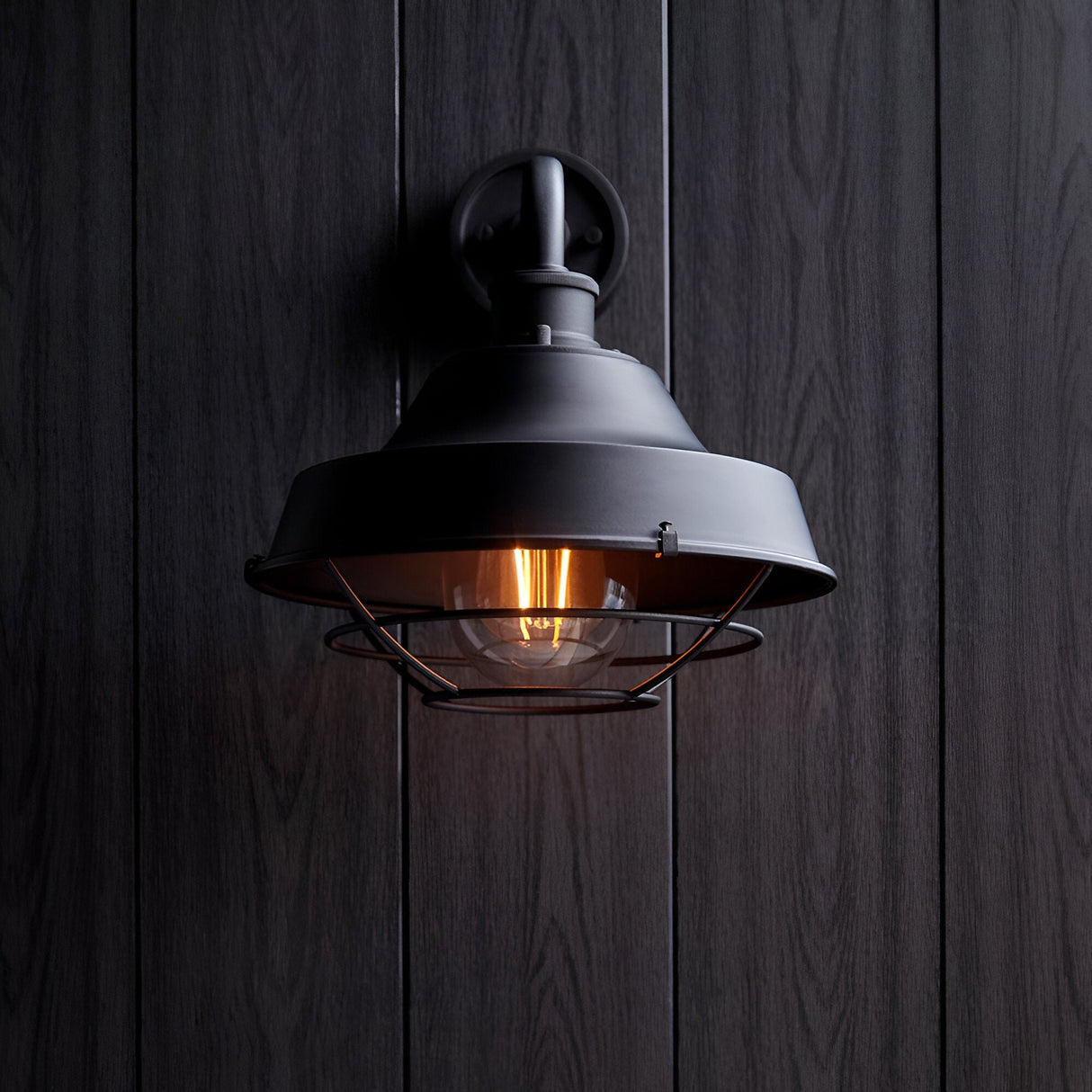 Caldton Outdoor Wall Light in matt black, industrial style with wire cage shade and exposed filament bulb, mounted on dark wood panelling.