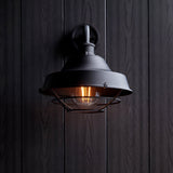 Caldton Outdoor Wall Light in matt black, industrial style with wire cage shade and exposed filament bulb, mounted on dark wood panelling.