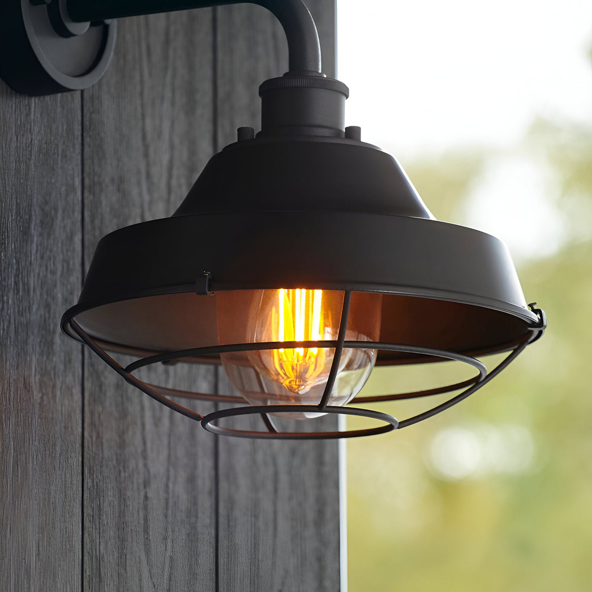 Caldton Outdoor Wall Light in matt black with die-cast body, wire cage shade and glowing filament bulb, mounted on a wooden panel outdoors.