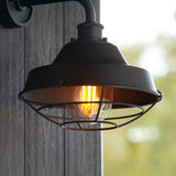 Caldton Outdoor Wall Light in matt black with die-cast body, wire cage shade and glowing filament bulb, mounted on a wooden panel outdoors.